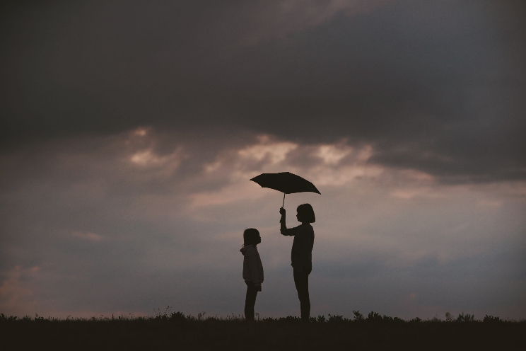 Adult holding an umbrella standing next to  a child on a cloudy evening.